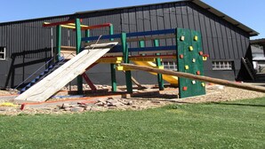 A playground with a slide, climbing wall, and swings, set against a building with a dark exterior. - Holiday Home in Burg Reuland (Burg Reuland)