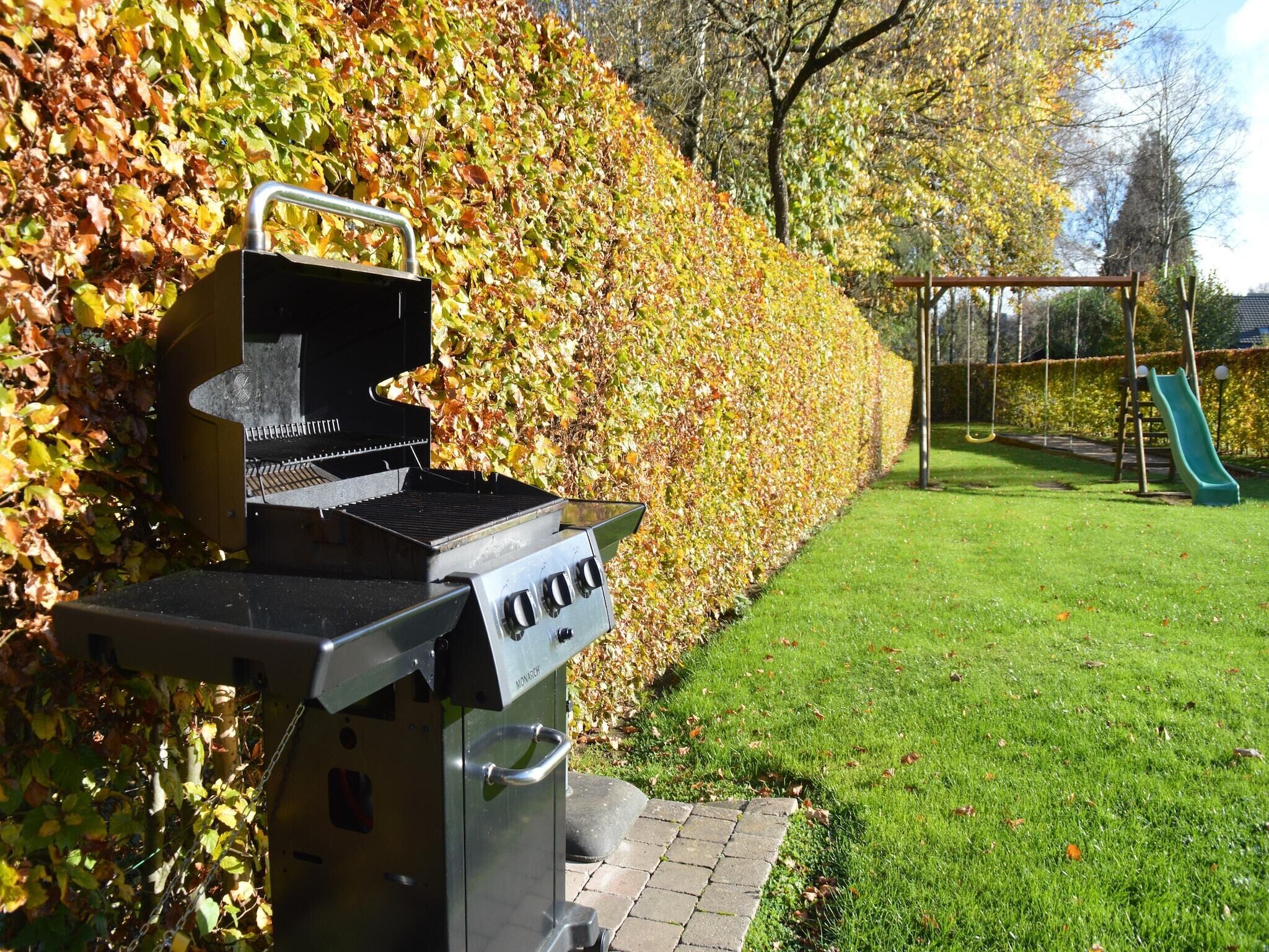 A barbecue grill in a garden with a hedge, a swing set, and a slide.