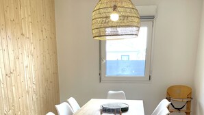 Un comedor con una mesa de madera, sillas blancas, una lámpara colgante tejida y una ventana con vistas.