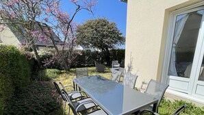 Un patio exterior con una mesa de vidrio y sillas de metal, rodeado de follaje y árboles en flor.