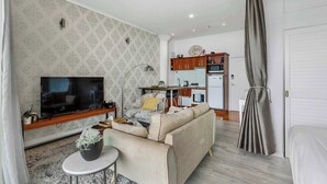 Ein Wohnzimmer mit einer Couch, einem Fernseher und einem kleinen Tisch mit einer Pflanze. Im Hintergrund ist eine Küche mit Schränken und einem Kühlschrank zu sehen.