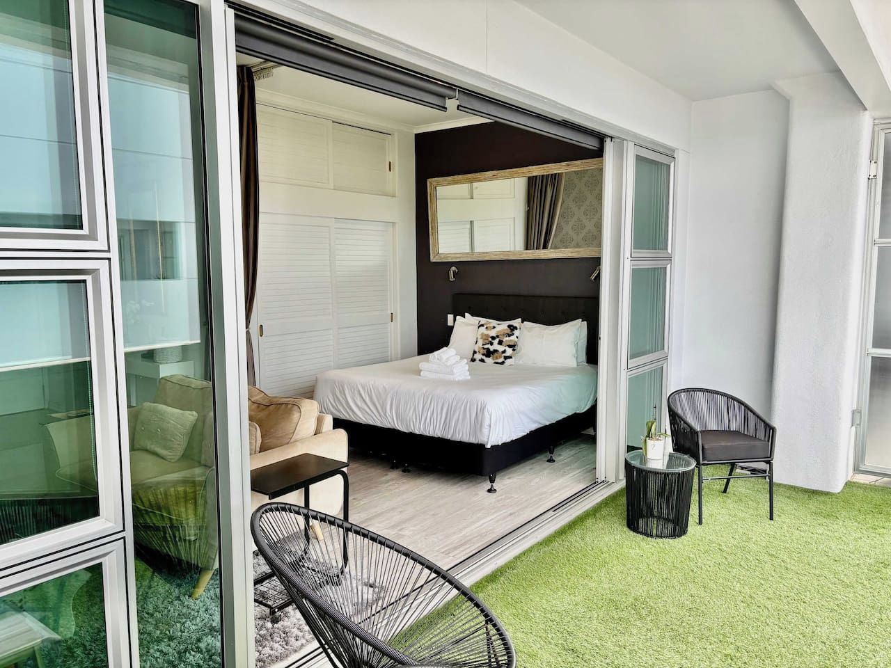 Ein Schlafzimmer mit einem Bett, einem Sessel, einem kleinen Tisch und einer Schiebetür, die auf einen Balkon mit künstlichem Rasen führt.