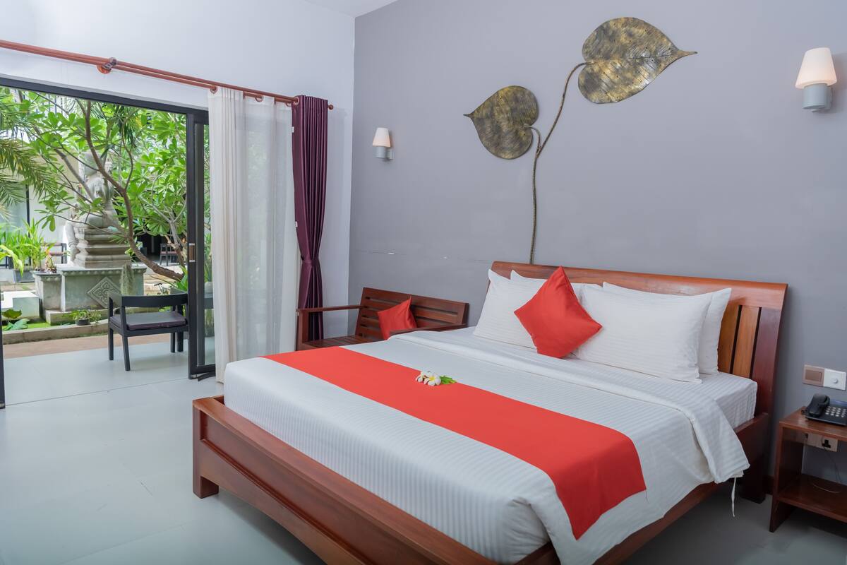 A hotel room with a bed, a nightstand, a chair, and a view of greenery outside.
