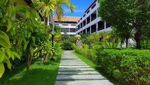 A pathway leading to a building with a statue, surrounded by lush greenery and palm trees. - New Riverside Hotel (Siem Reap)