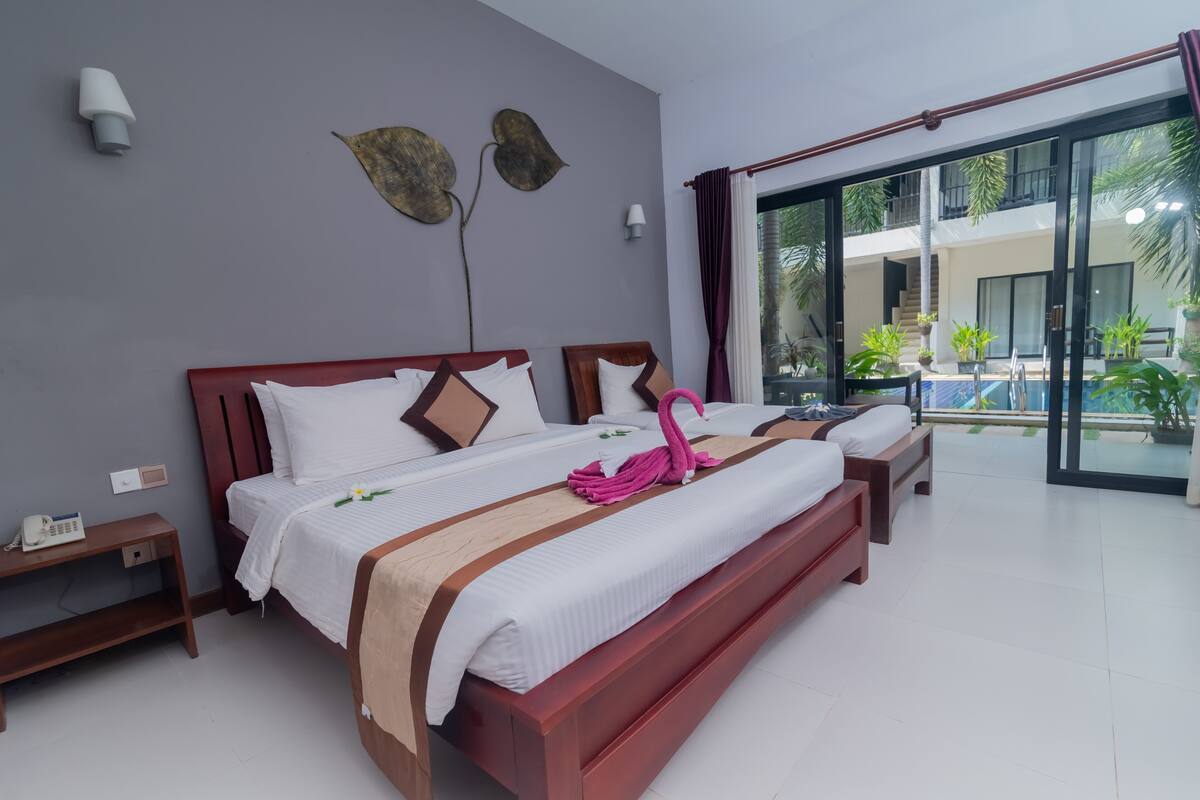 A hotel room with a large bed, a bedside table with a phone, and a view of a pool and greenery outside.