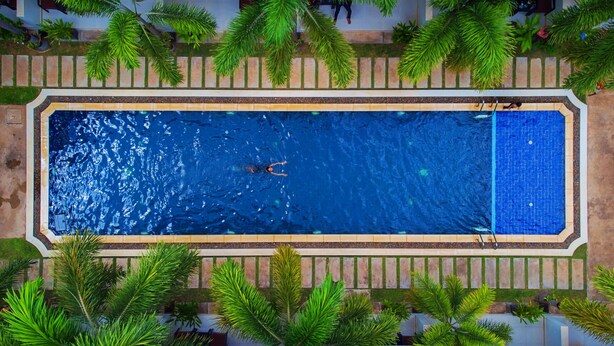 An aerial view of a rectangular swimming pool with two people swimming, surrounded by a tiled deck and palm trees. - New Riverside Hotel (Siem Reap)