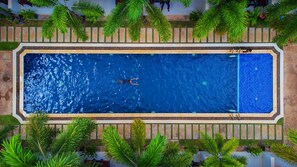 An aerial view of a rectangular swimming pool with two people swimming, surrounded by a tiled deck and palm trees. - New Riverside Hotel (Siem Reap)