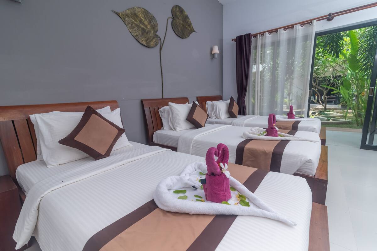 A hotel room with three beds, each with white bedding and brown pillows. There is a large window with a view of greenery outside.