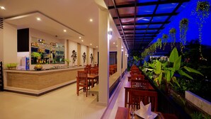 A restaurant with a bar area, wooden tables, and chairs. There are plants and a view of greenery outside. - New Riverside Hotel (Siem Reap)