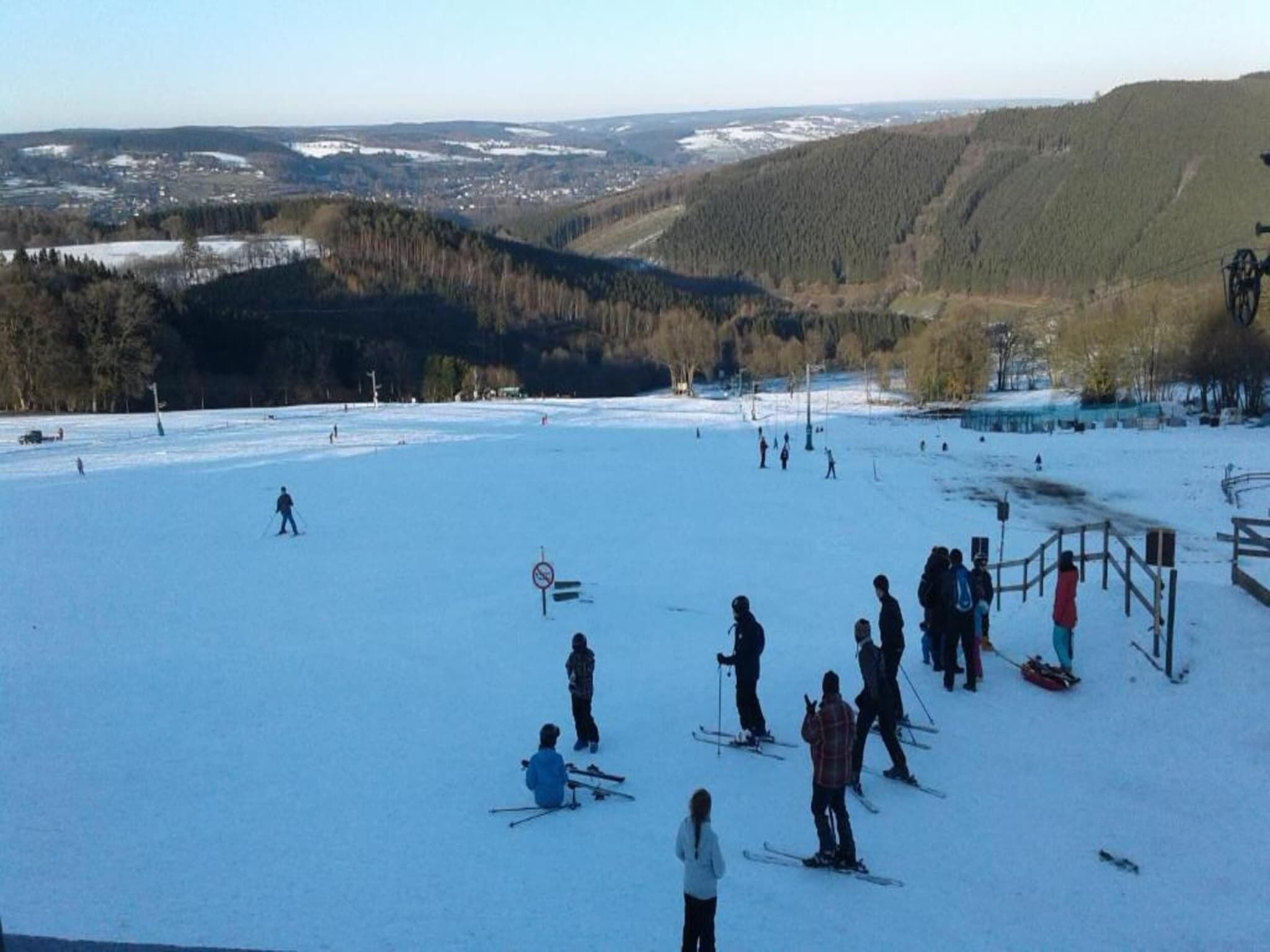 Una stazione sciistica con una vasta area innevata, impianti di risalita e una vista lontana di un paese e colline boscose.