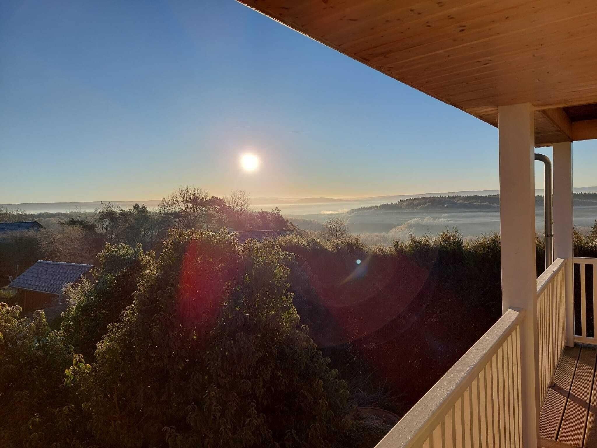 Un balcon en bois donnant sur un paysage où le ciel est dégagé et le soleil se trouve près de l’horizon.