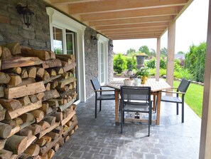 A covered patio with a stone wall, a wooden table, and chairs. - Luxury Ardennes chalet spa (Septon)