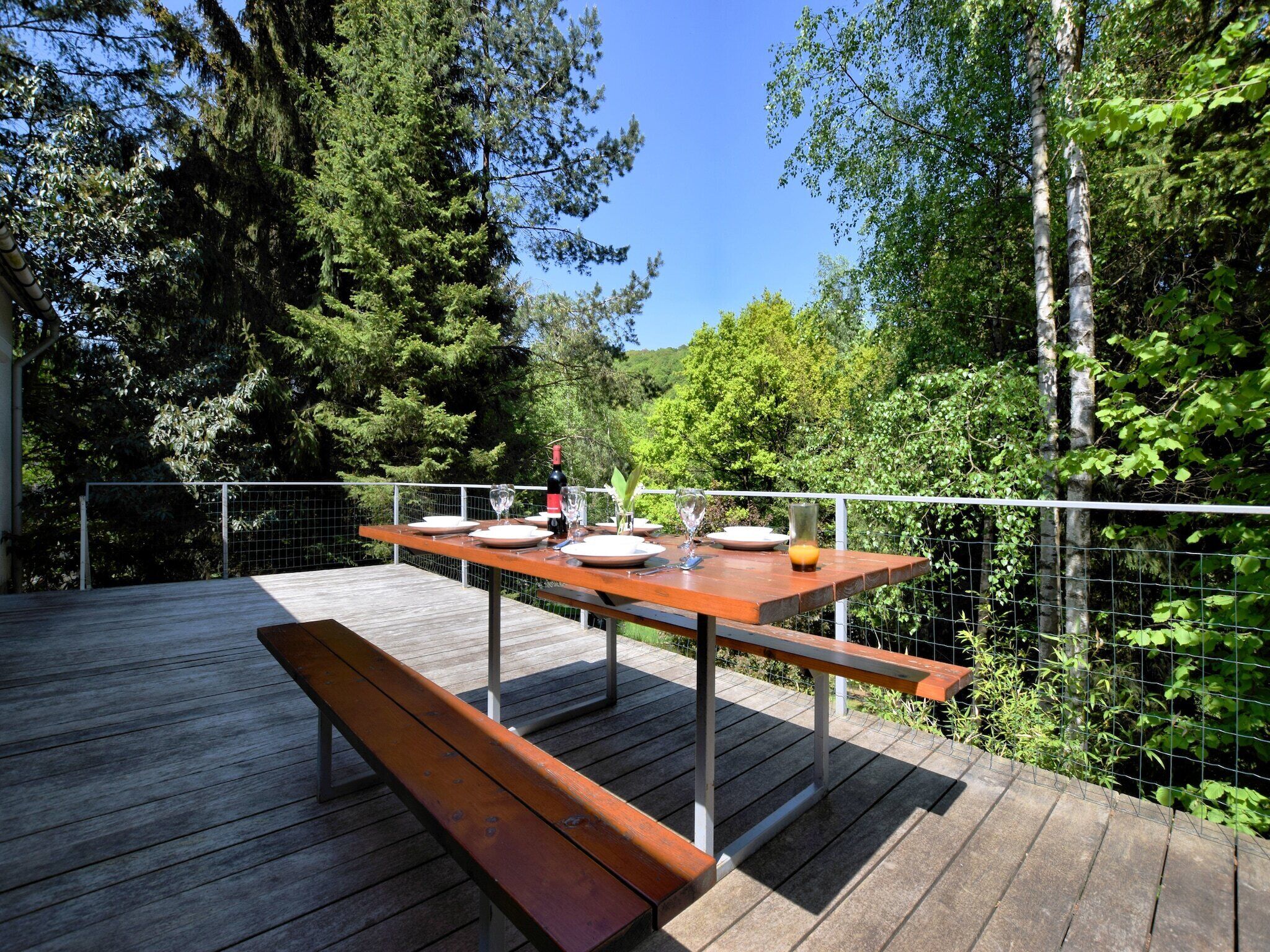 Una terrazza esterna in legno con un tavolo apparecchiato per mangiare, circondata da una recinzione metallica e vegetazione lussureggiante.