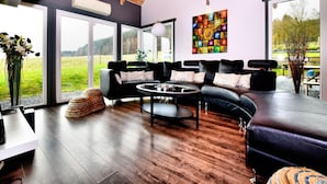 A modern living room with a wooden ceiling, large windows, a black leather sofa, a coffee table, and a colorful abstract painting. - Beautiful villa with heated outdoor pool (Manhay (lamormenil))