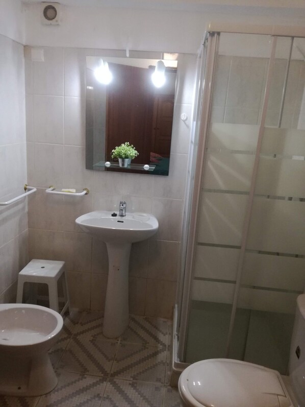A bathroom with a pedestal sink, a toilet, a small stool, a mirror with lights, and a shower enclosure.