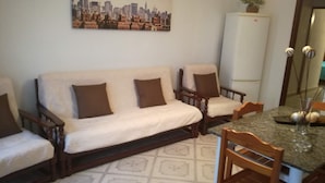 A living room with a sofa, two armchairs, a dining table with chairs, a refrigerator, and a cityscape painting on the wall.