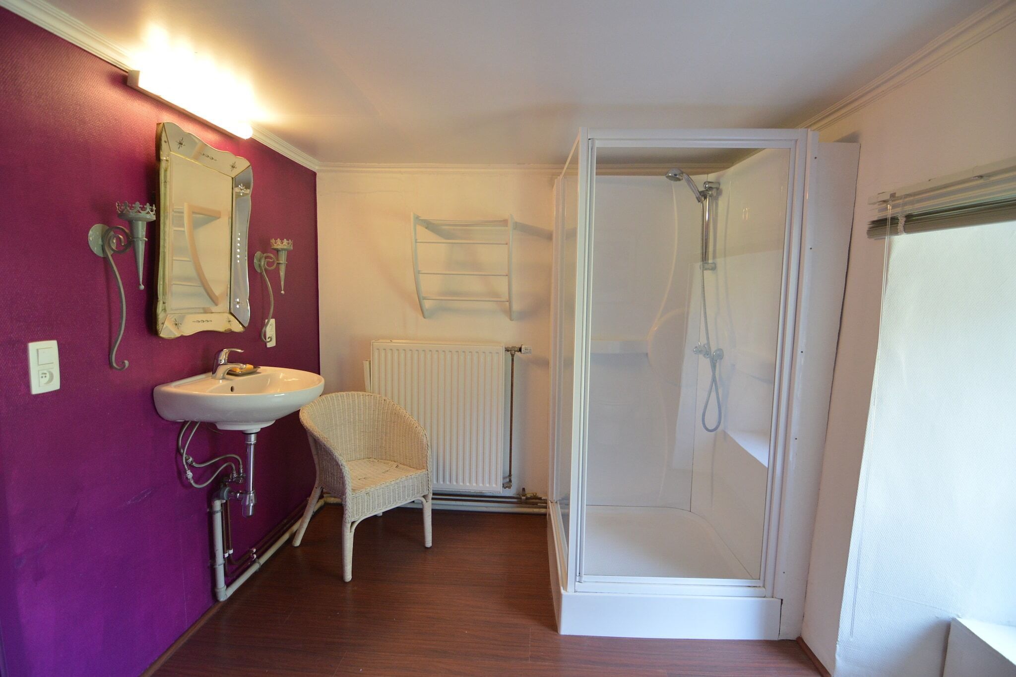Een badkamer met een wit wastafelmeubel, een rieten stoel, een douchecabine en een spiegel met decoratieve wandlampen.