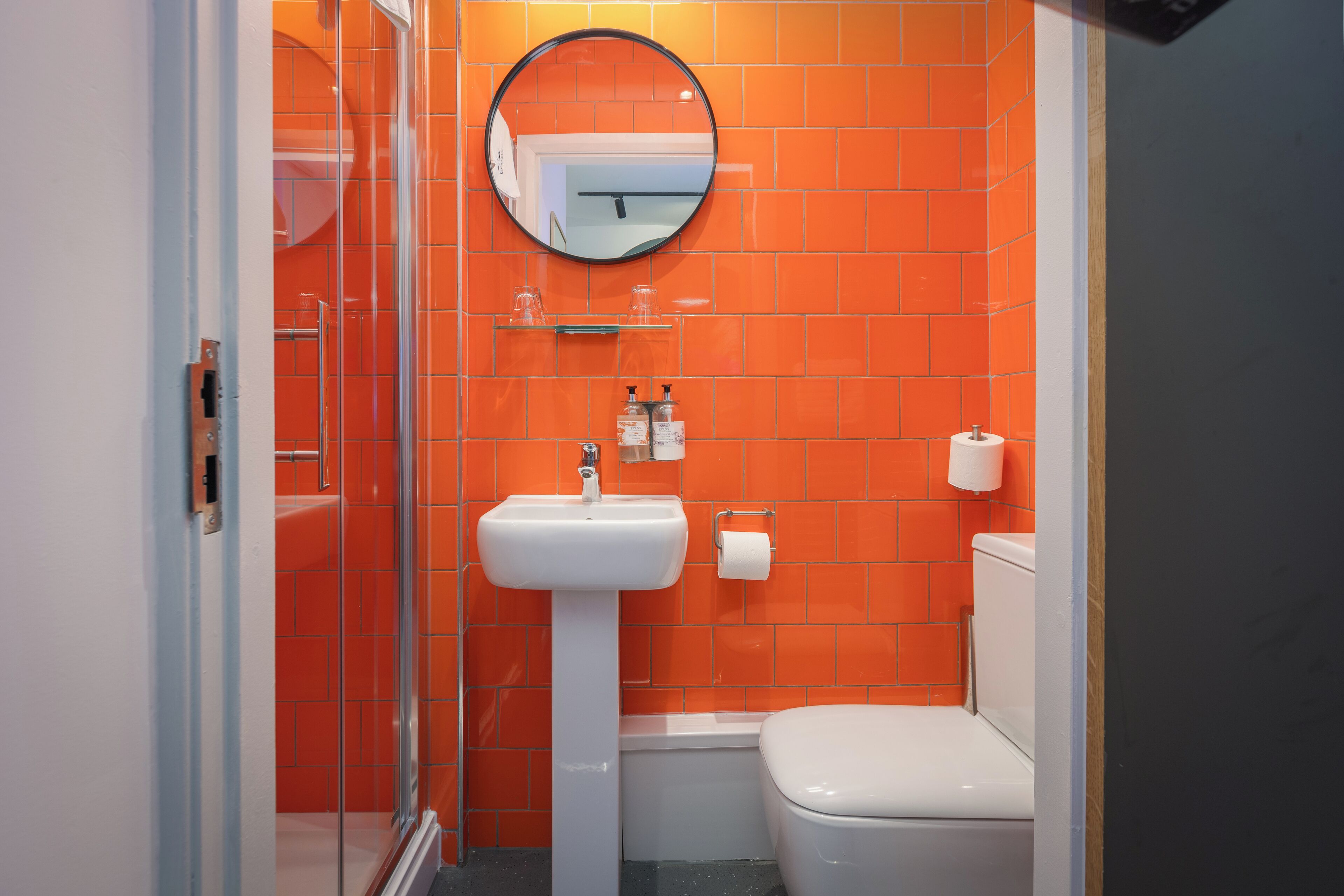 A bathroom with orange tiles, a white sink, a toilet, and a glass shower enclosure.