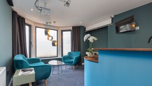 A modern hotel room with a curved reception desk, teal armchairs, a small round table, and a large window with curtains. - ART B&B (Blackpool)