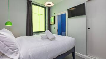 A neatly made bed with white linens and two rolled towels, a green pendant light, a window with curtains, a blue door, and a flat-screen TV mounted on the wall.
