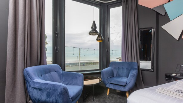 A modern hotel room with two blue armchairs, a small round table, a bed, and a large window offering a view of the city. - ART B&B (Blackpool)