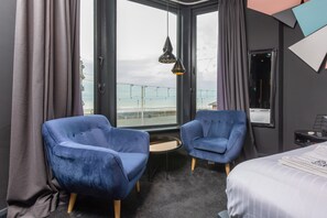 A modern hotel room with two blue armchairs, a small round table, a bed, and a large window offering a view of the city.