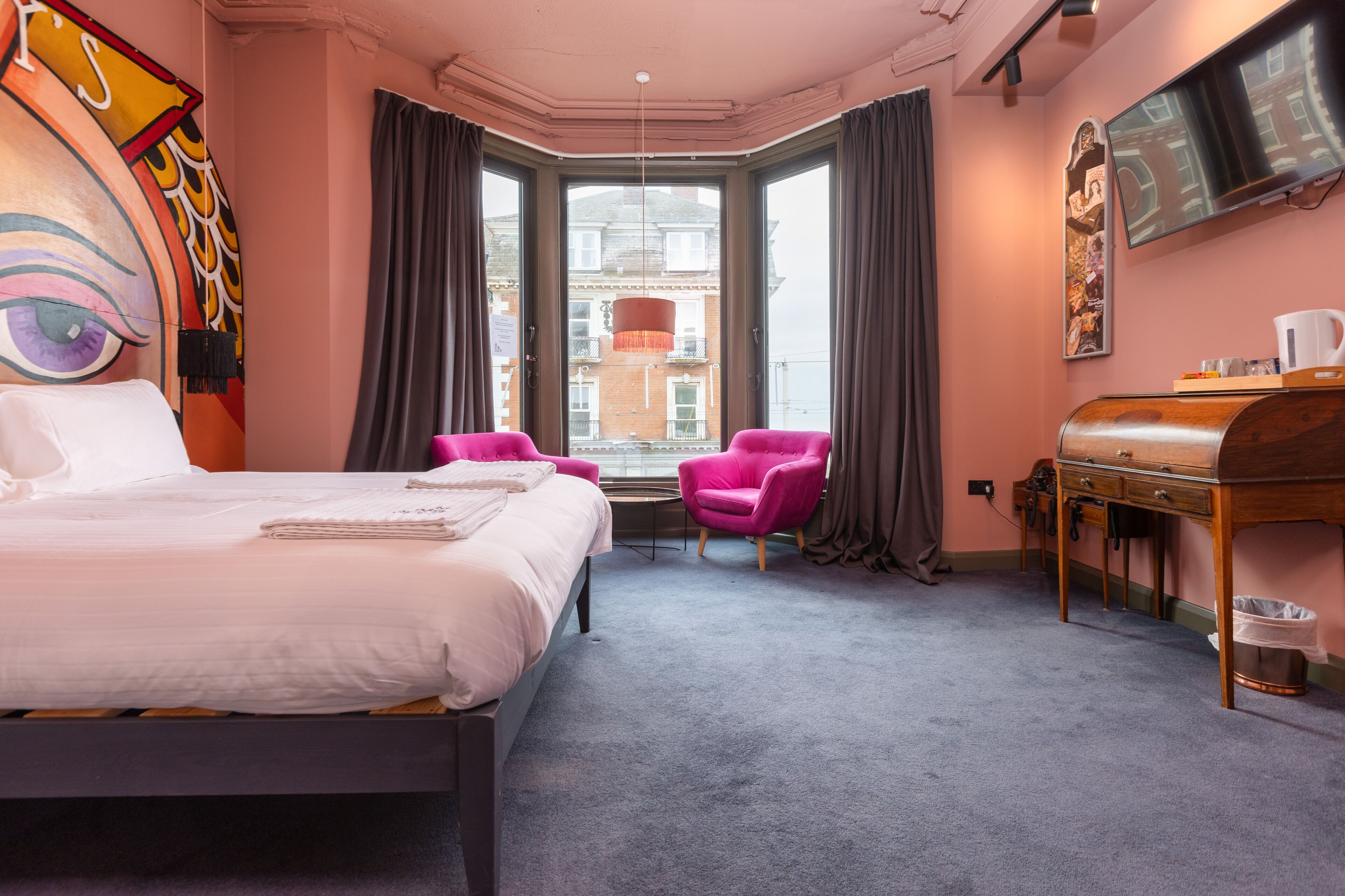 A hotel room with a large bed, two pink chairs, a desk, and a television.