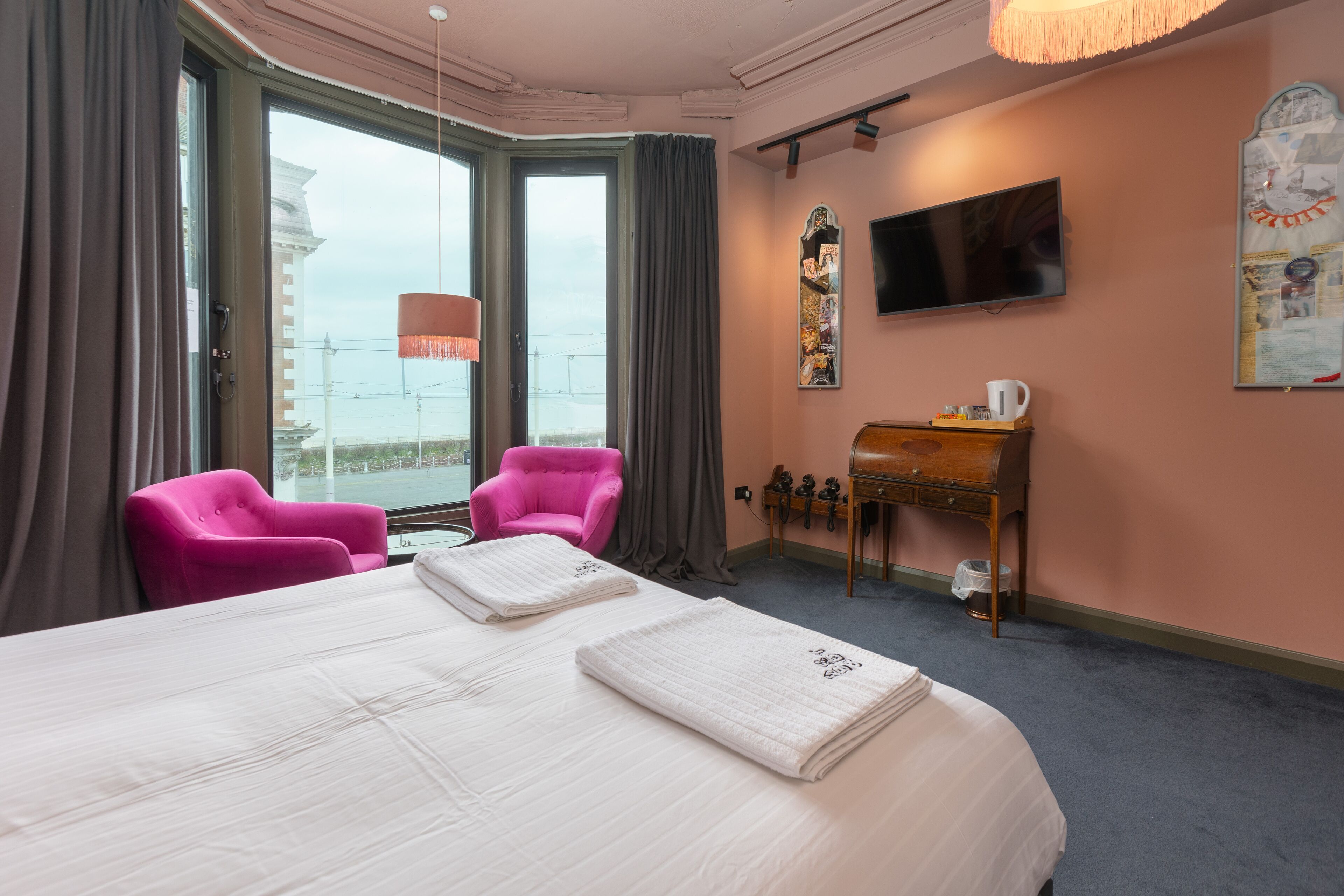 Habitación de hotel con una cama grande, dos sillas rosadas, un escritorio de madera, una televisión y vistas a un edificio a través de la ventana.
