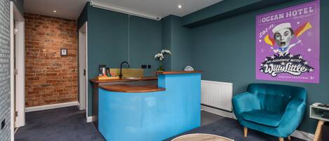 Un vestíbulo de hotel moderno con un mostrador de recepción azul, un sillón azul y una mesa redonda de madera.