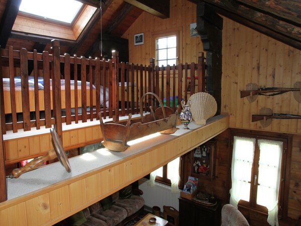 A loft bedroom with a sloped ceiling, wooden beams, a loft bed, a wooden chest, a decorative vase, and a shelf with books and a camera.