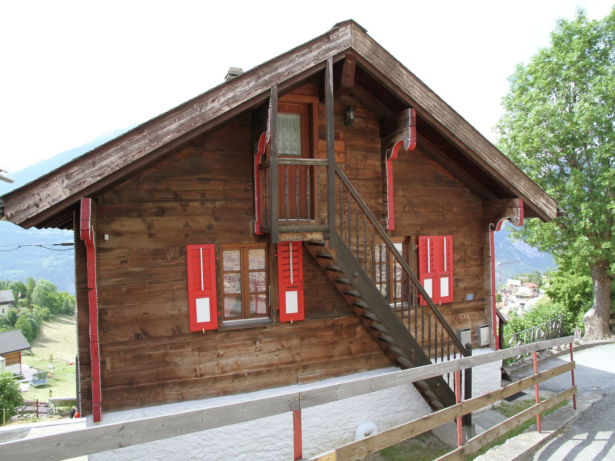 A wooden cabin with a wooden staircase leading to a balcony, surrounded by a fence and a view of the mountains.
