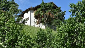 A traditional chalet with a sloped roof, wooden balconies, and a white facade, surrounded by lush greenery.