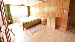 A bedroom with a wooden floor, a bed with a green bedspread, a bedside table, a wardrobe, and a window with curtains.