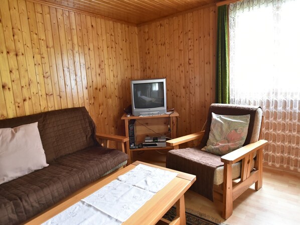 Una habitación con paneles de madera, un sofá marrón, un sillón de madera con un cojín floral y un televisor sobre un mueble de madera.