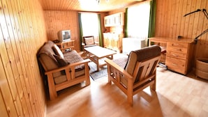 A cozy wooden cabin interior with a sofa, chairs, a small table, a TV, and a kitchenette.
