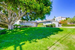 A residential house with a well-maintained lawn, a large tree in the foreground, and a clear blue sky.
