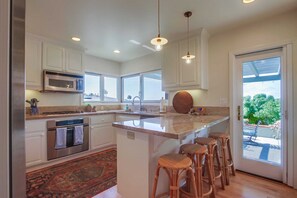 A modern kitchen with white cabinets, stainless steel appliances, a large island with bar stools, and a view of greenery outside.