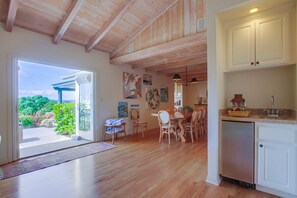 A spacious room with wooden floors, a dining area with chairs and a table, a kitchen with cabinets and a sink, and a view through a glass door to an outdoor area with greenery. - Solana Beach House w/ Hot Tub & Ocean Views (Solana Beach)