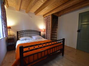 A bedroom with a wooden ceiling, a bed with an orange bedspread, a nightstand with a lamp, a staircase, and a green door.