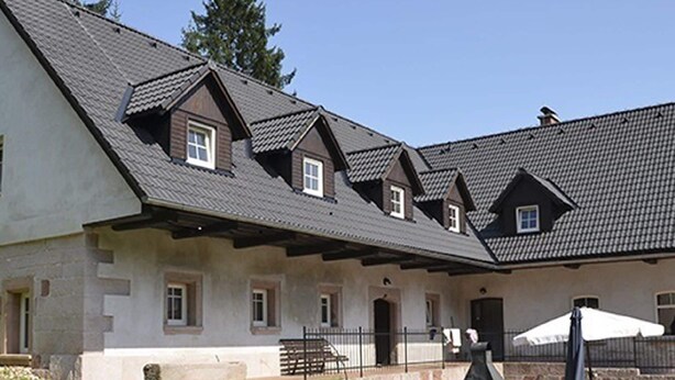 Un edificio con varios tejados a dos aguas, base de piedra y un pequeño porche con valla y mobiliario exterior.