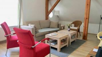 Un salon comprenant un canapé, un fauteuil rouge, une table basse en bois et une chaise en osier.