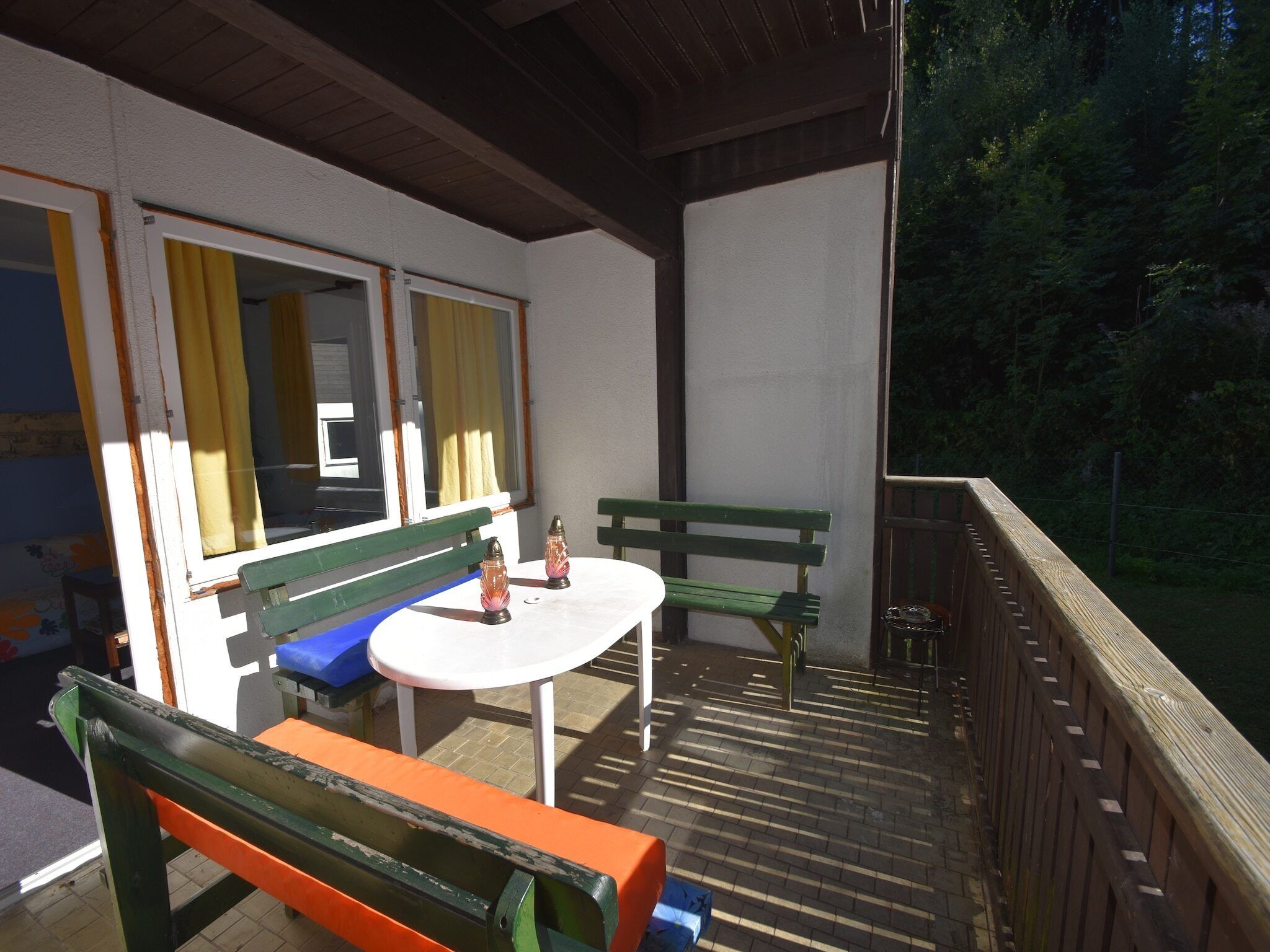 Un espace extérieur abrité, avec une table et des bancs, une fenêtre avec des rideaux et une vue sur une zone boisée.