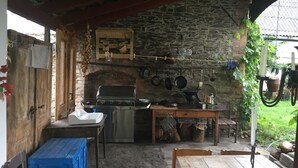 Une cuisine d’extérieur rustique, avec un mur en pierre, un barbecue, des ustensiles de cuisine suspendus et une table en bois.