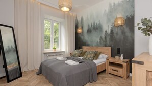 A bedroom with a bed, a nightstand, a window with curtains, a wall mural, and a hanging light fixture.