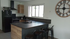 A modern kitchen with a central island, black appliances, and a large wooden clock on the wall. - Maison Neuve de 2023, Plain Pied, 2 Chambres, à 10 Minutes à Pied de la Plage (Fréhel)
