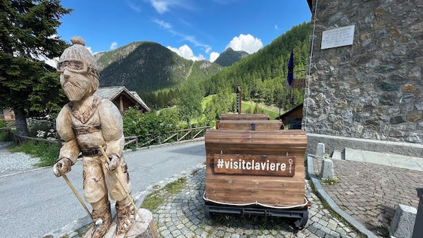 Una estatua de madera de un esquiador, un letrero de madera con #visitclaviere, y un edificio de piedra con un cartel.
