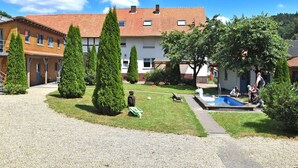 Une cour avec une piscine, entourée de bâtiments aux toits recouverts de tuiles rouges et d’arbres aux branches soigneusement taillées.