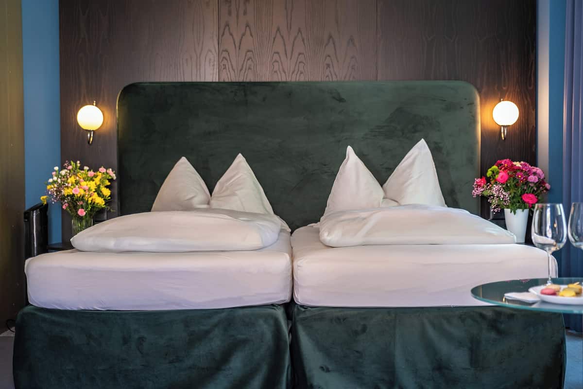 A double bed with a green upholstered headboard, two bedside tables with lamps, a vase of flowers, and a glass table with a plate and a wine glass.