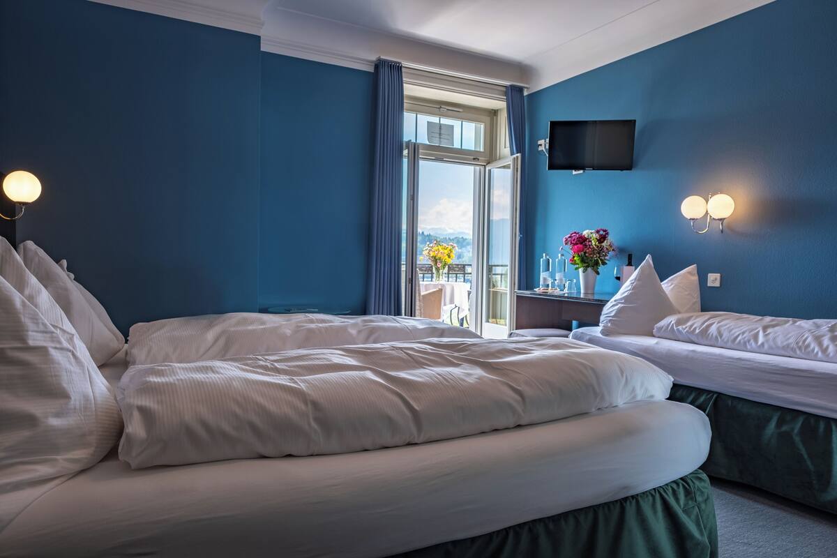 A hotel room with two beds, a TV mounted on the wall, a window with curtains, and a view of the outdoors.