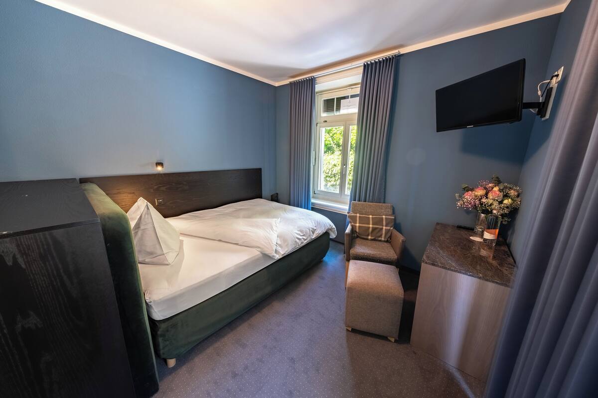 A bedroom with a bed, a desk, a chair, a TV, and a window with curtains.